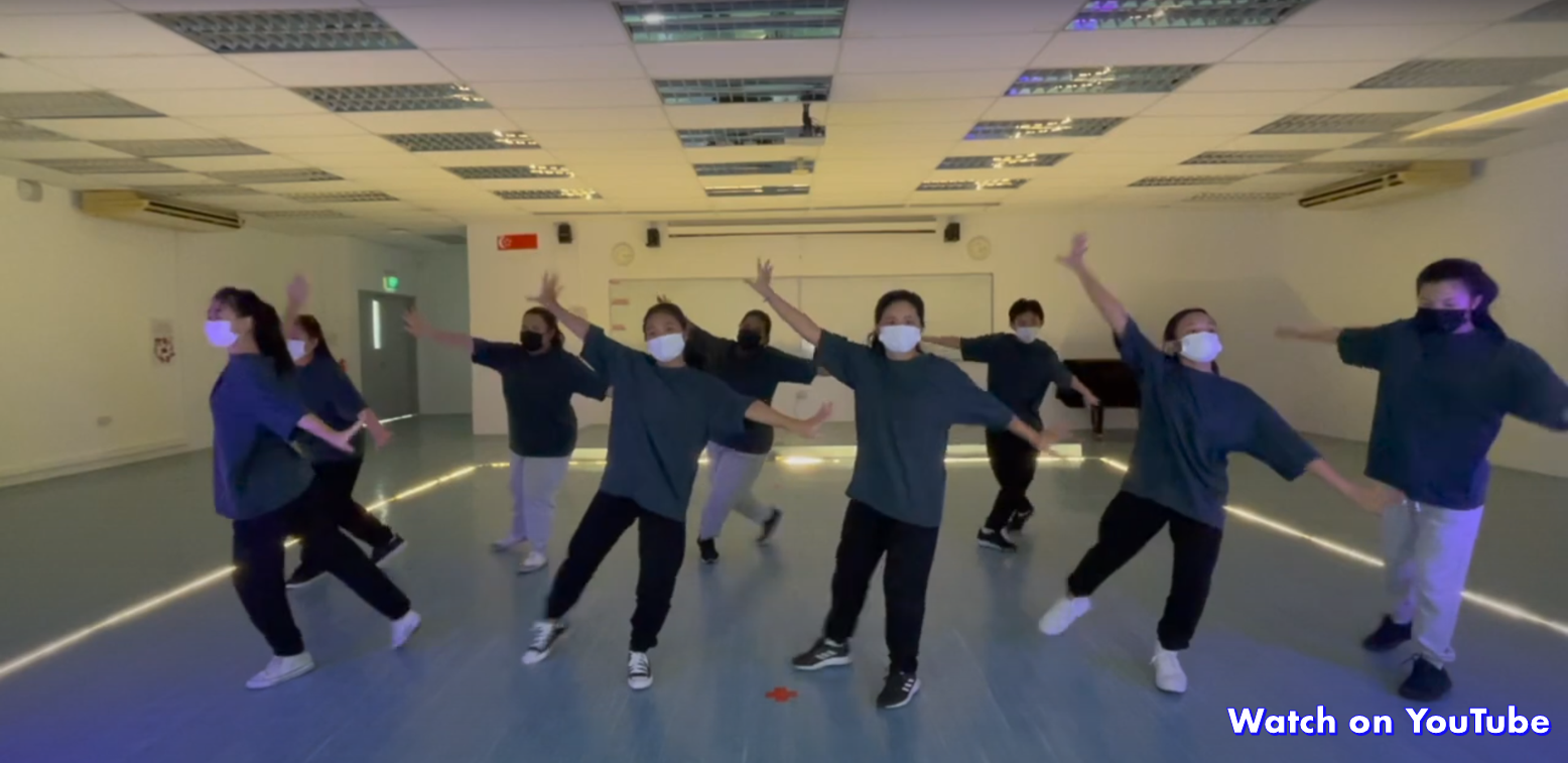 A group of masked dancers in dark clothing perform synchronized moves in an indoor studio with tiled ceiling.